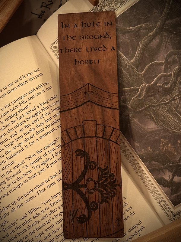 Walnut bookmark engraved with 'In a hole in the ground there lived a hobbit' above a round hobbit door, resting on an open copy of The Hobbit
