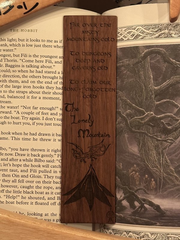 Walnut bookmark engraved with 'Far over the misty mountains cold', the words 'The Lonely Mountain', a dragon and a mountain peak, resting on an open copy of The Hobbit